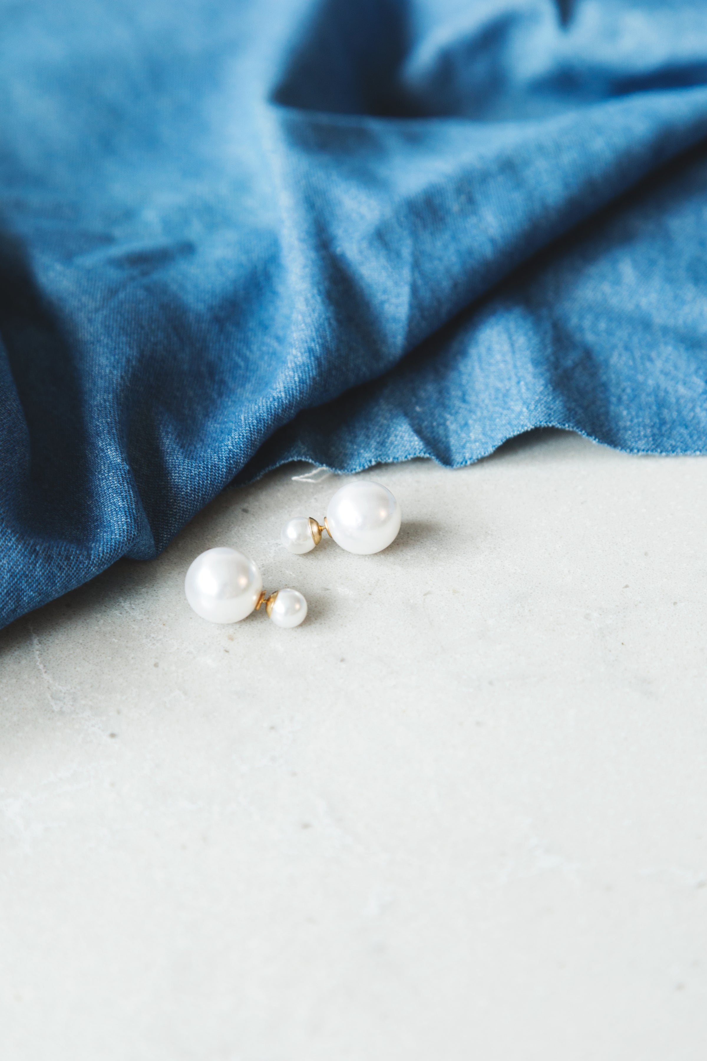 Elegant double-pearl stud earrings placed on a white surface with a blue fabric background.