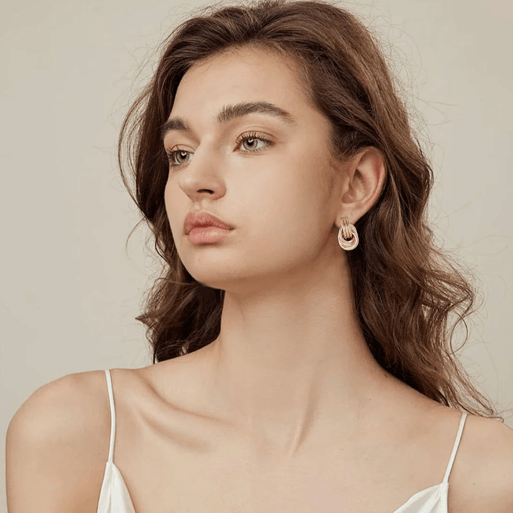 A woman with wavy brown hair wearing gold-tone stacked hoop earrings, styled with a minimalist white outfit, showcasing a classic and elegant look.