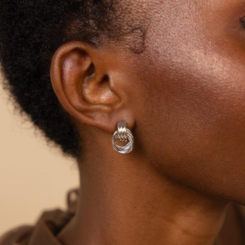 A close-up of a woman wearing silver-tone interlocking hoop earrings, styled with natural hair and a neutral-toned outfit, emphasizing the earrings' sleek and modern design.