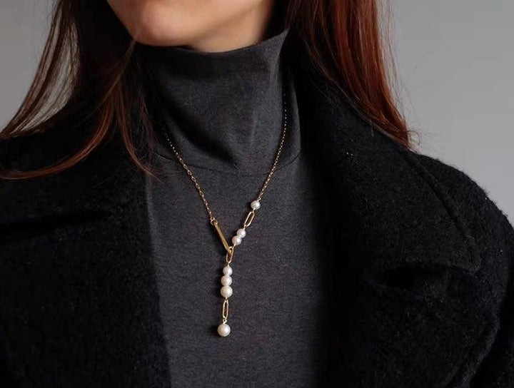 Gold chain necklace with white pearl accents worn over a grey turtleneck and black coat.