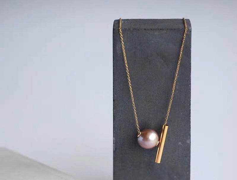 Gold chain necklace with a pink pearl and cylindrical gold bar pendant displayed on a grey stand.