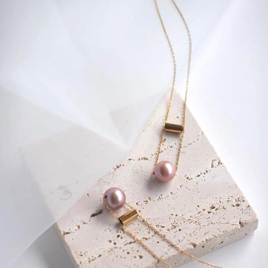 Delicate gold chain necklace with pink pearls and cylindrical gold accents on a textured surface.