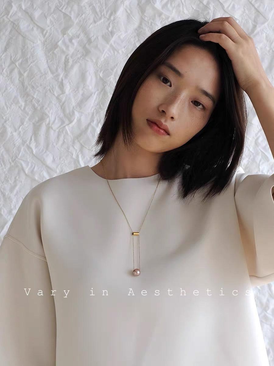 Minimalist gold necklace with a pink pearl pendant worn over a white oversized top.