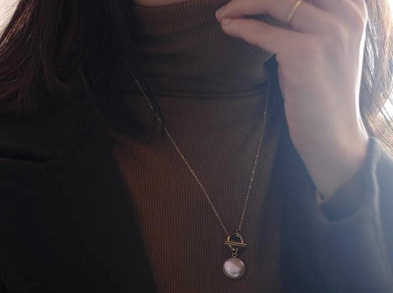 Gold toggle clasp necklace with a white pearl pendant worn over a brown turtleneck sweater.