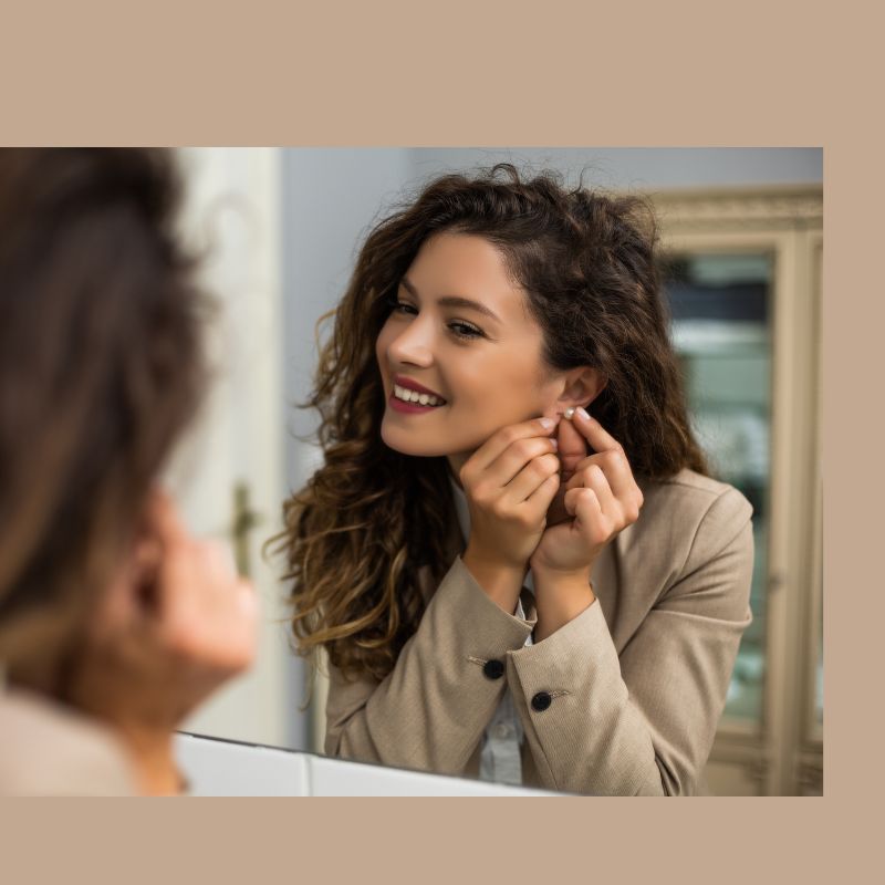 Smiling woman in a beige blazer putting on pearl earrings while looking in the mirror.